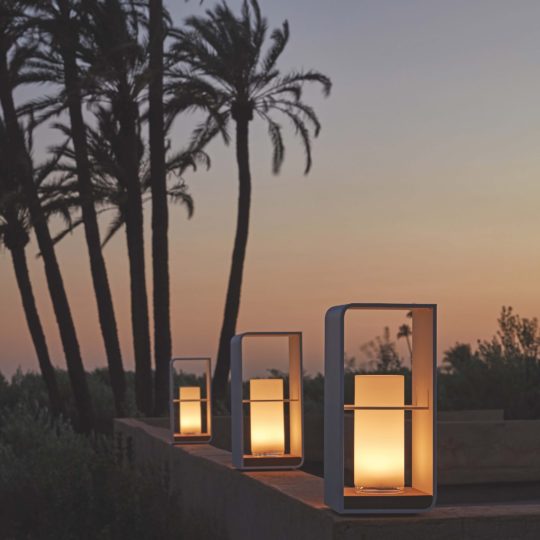 The Contemporary Outdoor Designer LED Lantern Set casts a warm glow on a ledge at sunset, framed by tall palm tree silhouettes and a vibrant sky for a peaceful, modern outdoor scene.