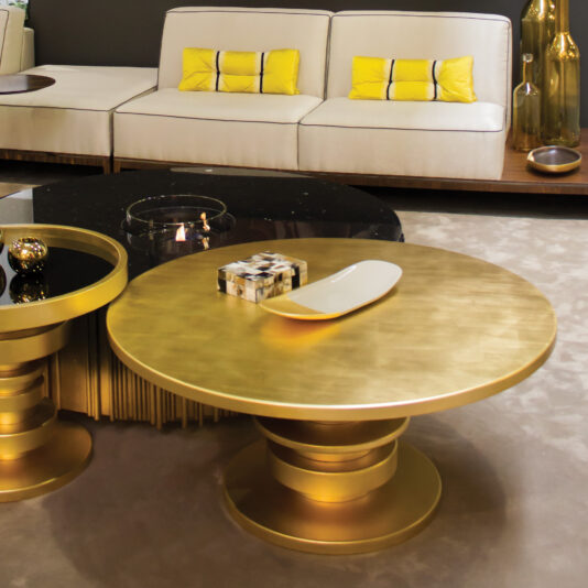 A modern living room with a white sofa, yellow and black cushions, a striking designer round coffee table, glass candle holder, decorative objects, and gold vases, all arranged on a beige carpet.