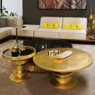 A modern living room with a cream sofa and yellow-accented cushions, two gold coffee tables, the Striking Designer Round Side Table, decorative objects, and gold vases—all against dark walls and bold contemporary art.