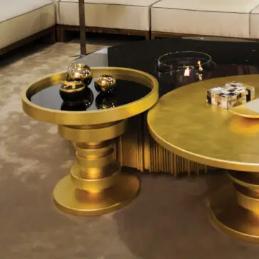 Modern living room with a beige sectional and two round, gold pedestal coffee tables. The Striking Designer Round Side Table holds golden spheres, whilst another table displays a glass bowl and book on the beige carpeted floor.
