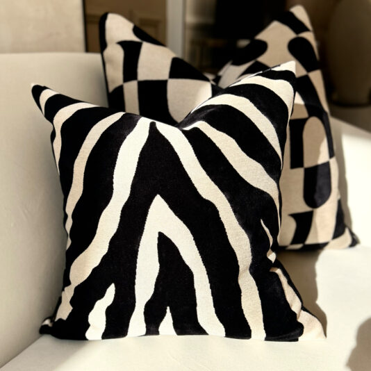 The Luxury Velvet Zebra Pattern Scatter Cushion graces a light-colored sofa alongside another pillow featuring black rectangles on white, both basking in sunlight to enhance the refined charm.