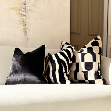 Luxury Velvet Zebra Pattern Scatter Cushion The white sofa is adorned with three luxury velvet scatter cushions: a plain black one, the elegant Luxury Velvet Zebra Pattern Scatter Cushion, and one featuring a striking geometric design. A textured wall with abstract art enhances the scene's sophistication.
