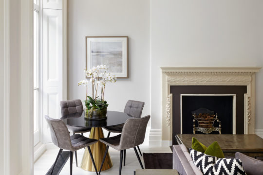 A modern living room with a round black dining table and four cushioned chairs. The table features an orchid centerpiece. A decorative fireplace to the right showcases a framed landscape painting above it. As part of this interior design project, the room boasts a neutral color scheme and large windows.