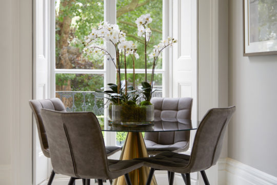 A modern dining area featuring a round glass table with a gold base and four gray upholstered chairs. As part of this interior design project, a clear vase with white orchids serves as a centerpiece. The room has large windows offering views of the greenery-filled yard outside.