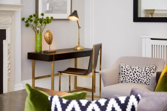 A stylish living room, part of a recent interior design project, features a small, dark wood desk with gold legs, adorned with a green vase of flowers, a modern art piece, and a brass desk lamp. A contemporary black chair sits by the desk. The room also boasts a light grey armchair with a black-and-white polka dot pillow.