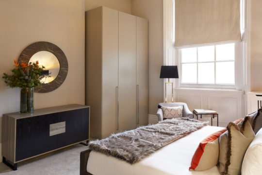 A modern bedroom, part of an exquisite interior design project, features a neatly made bed with a fur throw and decorative pillows. Beside the bed is a beige armchair, a black floor lamp, and a small table. A large wardrobe stands against the wall, while a round mirror with a floral arrangement adorns a side cabinet.