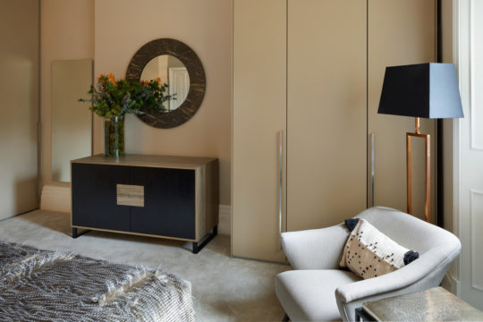 This interior design project features a modern bedroom with a large beige wardrobe, a round mirror above a wooden dresser adorned with a decorative floral arrangement, and an elegant armchair with a black and white patterned pillow. A floor lamp with a black shade is positioned near the window.