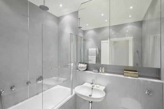 Modern bathroom with light gray walls featuring a white bathtub and glass shower partition, a small white sink with a mirror above, and a countertop with neatly arranged toiletries. As part of an interior design project, ceiling lights provide bright illumination in the space.