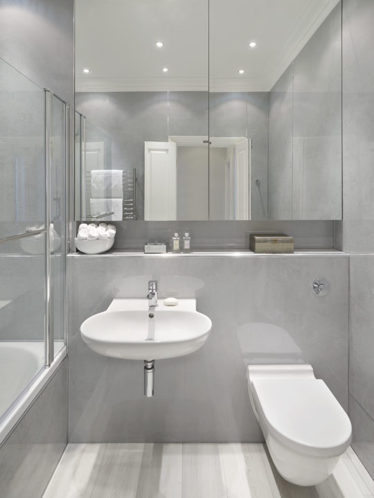A modern bathroom with a minimalist design, inspired by Knightsbridge interior design, featuring a wall-mounted sink with a mirror above, a toilet, a glass-enclosed shower, and a shelf with toiletries. The room has light grey walls, wood-textured flooring, and recessed ceiling lights.