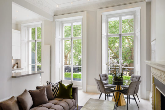 A modern, bright living and dining area with large floor-to-ceiling windows letting in natural light. This interior design project features a gray couch with cushions, a small dining table with four chairs, and a minimalist kitchen counter. Green foliage is visible outside the windows.