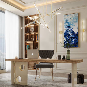Modern office space featuring a large wooden desk with a sleek design, an empty chair, a laptop, and books. The room is illuminated by an Abstract Gold Plated Contemporary Chandelier and natural light from a large window. A colorful abstract painting and built-in bookshelf adorn the background, adding to the elegance.