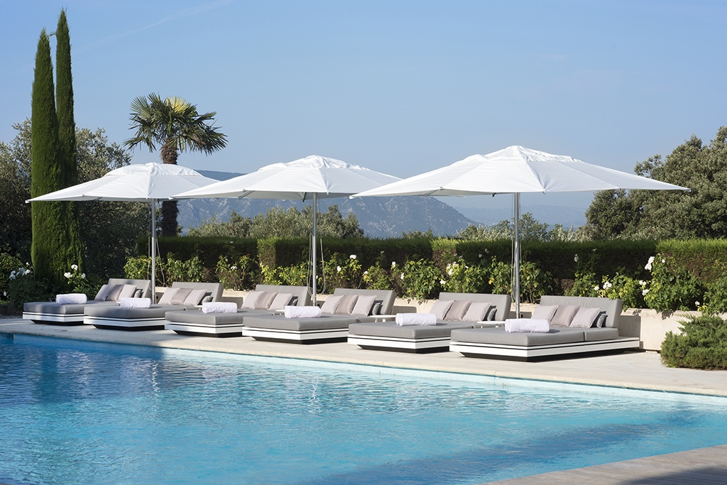 Several modern poolside lounge chairs with white cushions and large white umbrellas are arranged in a row next to a clear blue swimming pool. The backdrop includes lush greenery, tall trees, and distant mountains under a clear sky.