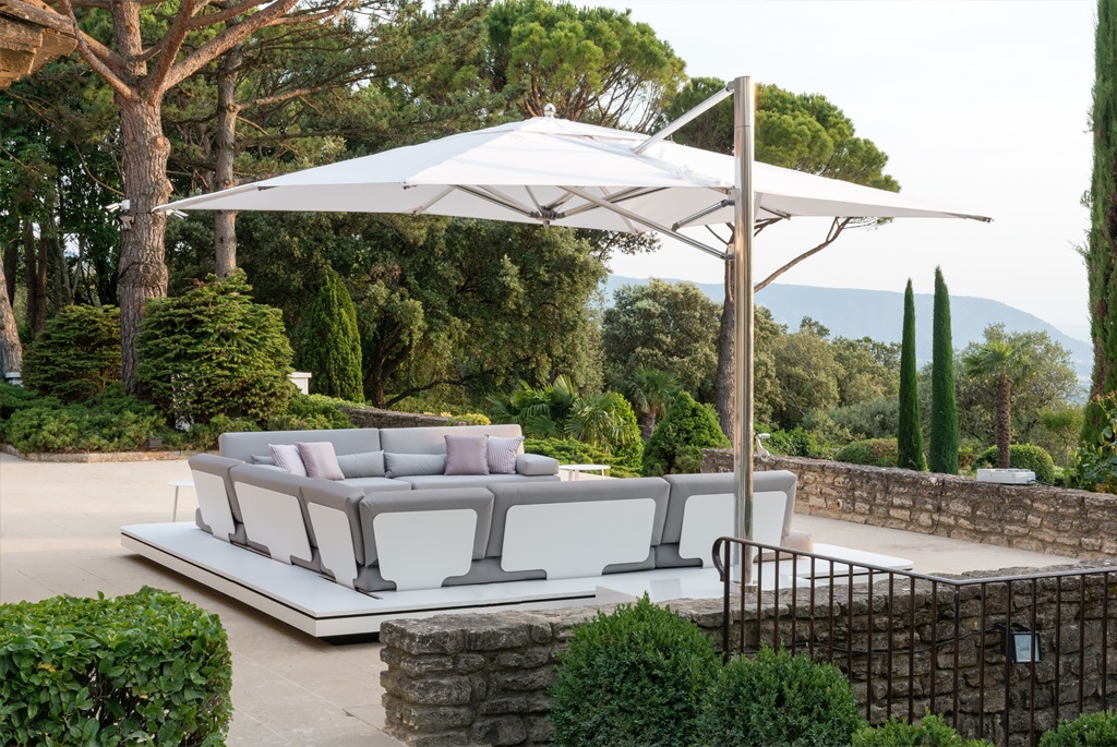 A modern outdoor seating area with a large white parasol covering gray and white modular sofas. Set on a spacious patio overlooking lush greenery, trees, and distant hills, it exudes a serene and luxurious ambiance.