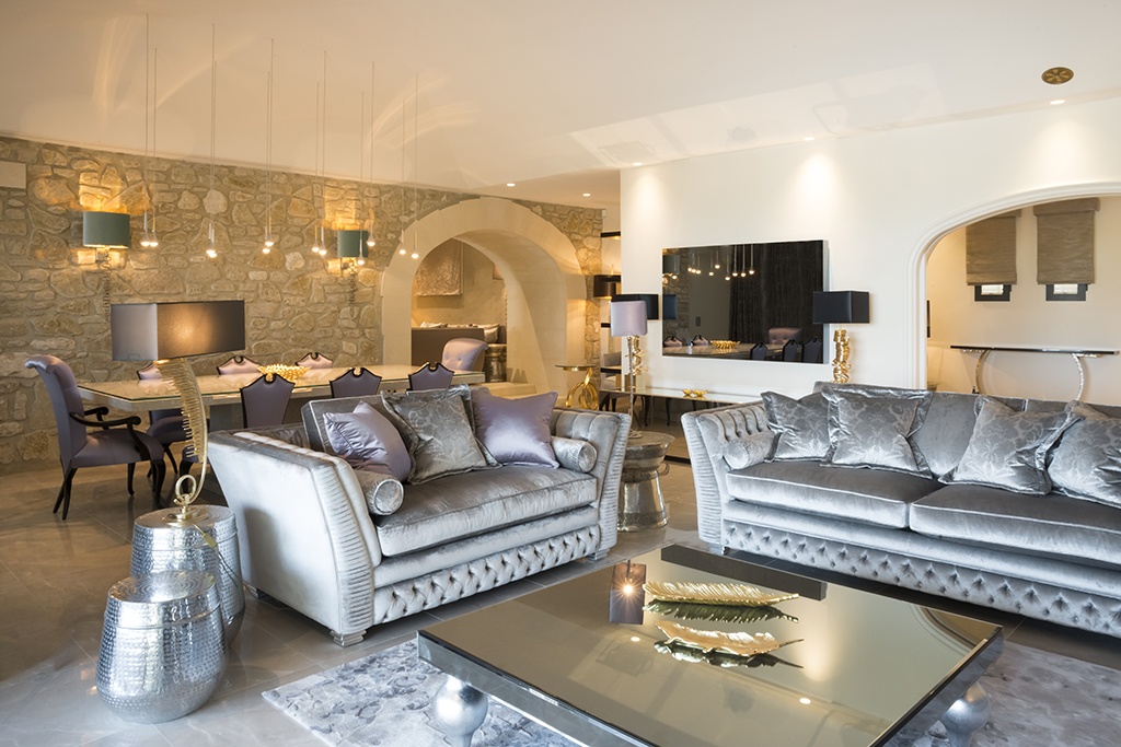 Modern living room with stone accent wall. Silver velvet sofas face a mirrored coffee table, adorned with decorative items. Open archways lead to a dining area with plush purple chairs and pendant lighting. The space is subtly lit with a sophisticated ambiance.