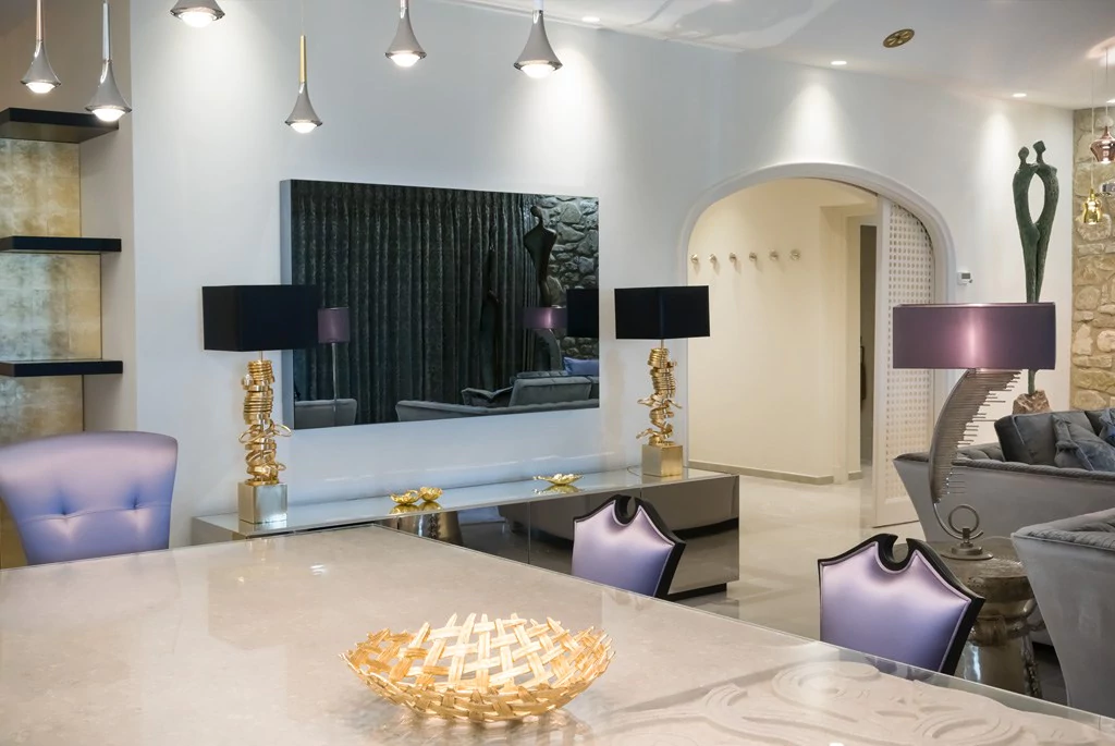 A modern living room with contemporary decor features stylish furniture, including a gray couch and purple chairs. Unique gold and black table lamps adorn an elegant table. A large wall mirror and decorative art pieces enhance the space's sophisticated ambiance.