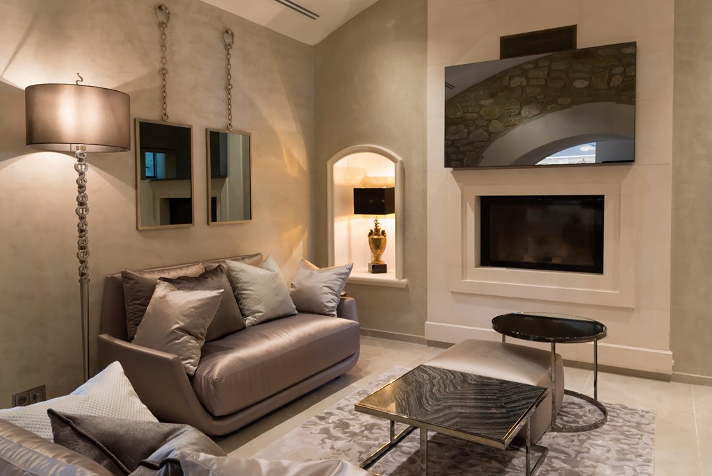 A modern living room with light beige walls, a comfortable sofa adorned with plush pillows, two mirrored wall accents, a floor lamp, and a marble coffee table. The room also features a built-in fireplace and decorative niche, creating a cozy yet elegant atmosphere.