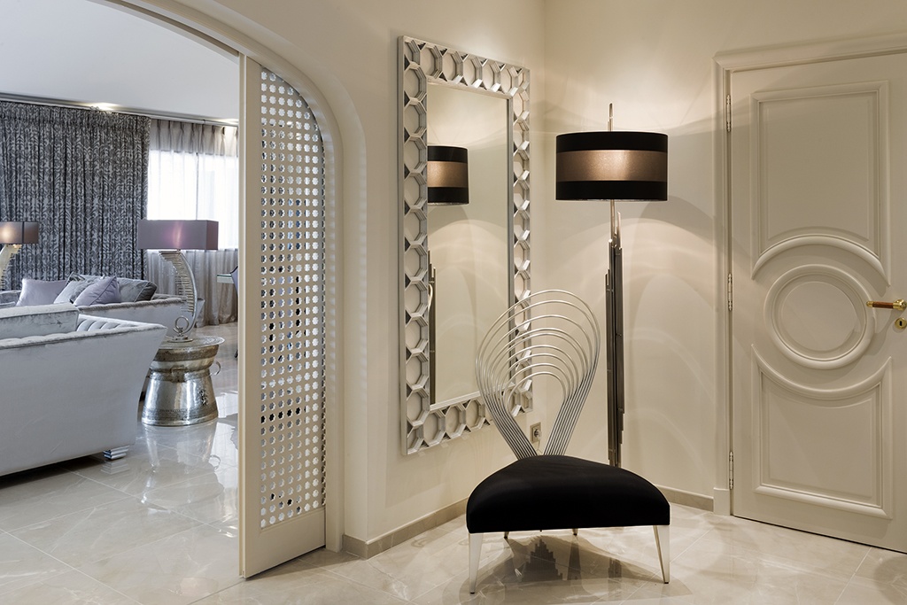 Modern indoor setting with a sleek black chair featuring a unique metallic backrest design, positioned near a large mirror with a patterned frame. A floor lamp stands beside the chair. A patterned archway leads to a cozy living room with gray seating and curtains.