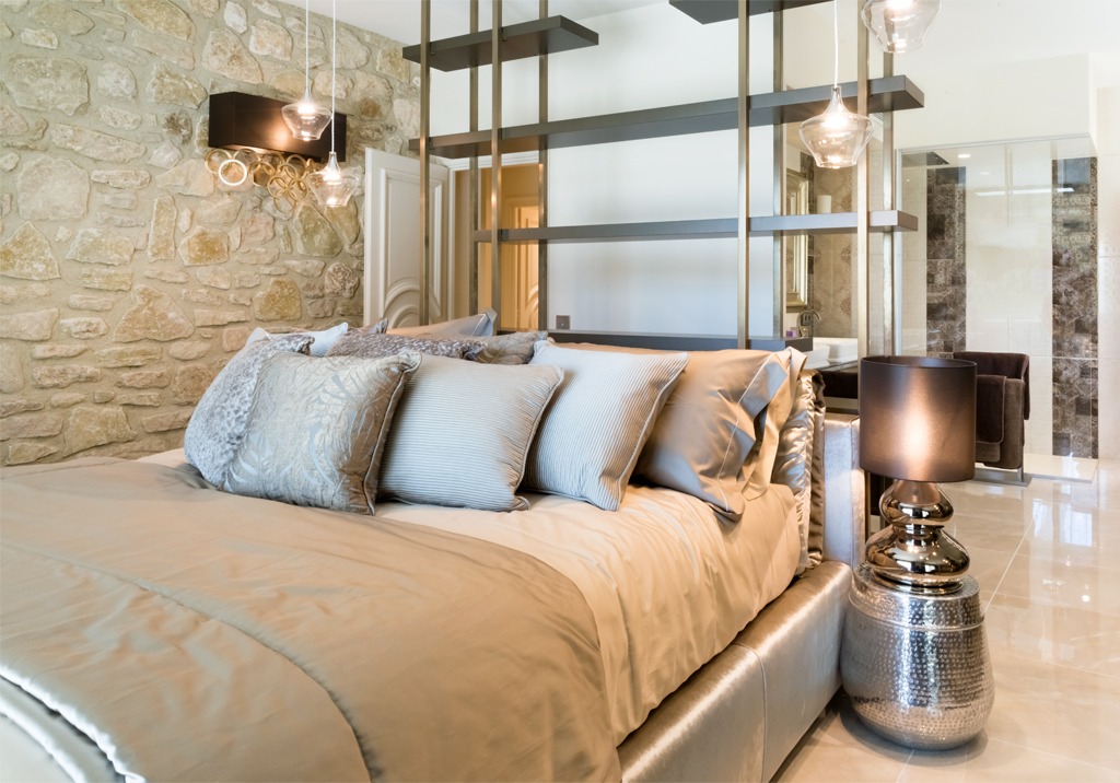 Modern bedroom with a stone accent wall, luxurious bedding, and metallic accents. A large bed with plush pillows and blankets is in the center, with a sleek dark bedside lamp on a metallic side table. Elegant pendant lights hang from the ceiling above the bed.