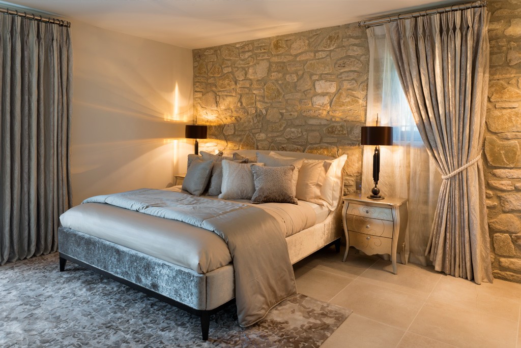 A cozy bedroom with a stone accent wall and two bedside tables featuring black lamps. The bed is adorned with beige linens and pillows. The walls are light-colored, and floor-length grey curtains frame the windows. A light gray rug covers the floor.