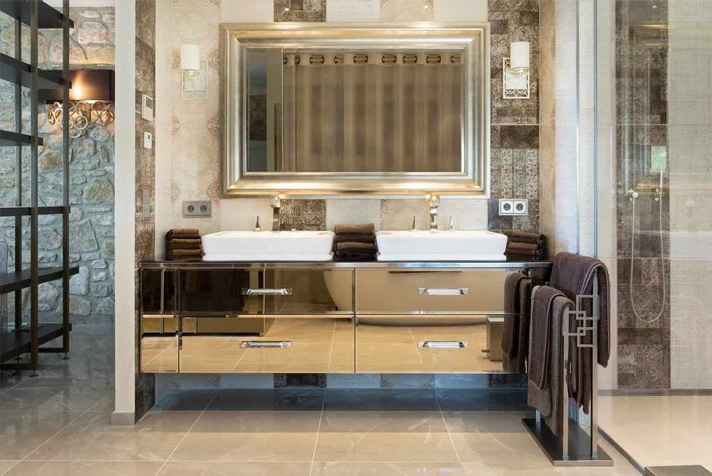 A modern bathroom features a large mirror with a gold frame above a double sink vanity unit. The vanity has a sleek, mirrored facade and is flanked by dark towels. To the right is a glass-enclosed shower. The space has tile flooring and walls with a mix of patterns.