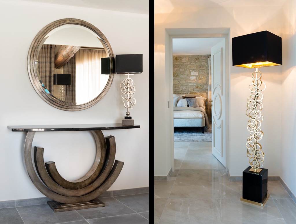 The image shows two views of a modern, stylish interior. The left view features a round mirror above an art deco console table with a sculptural base and a black lamp. The right view shows a hallway with a tall, ornate floor lamp, leading into a bedroom with a stone wall.