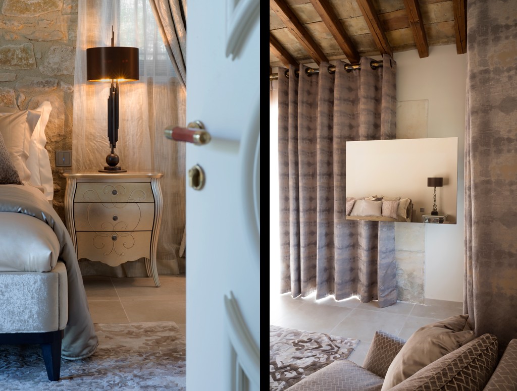 A cozy bedroom features a plush bed next to a wooden nightstand with an elegant lamp. A white door is slightly ajar. The adjoining room has tall gray curtains, a large wall mirror, and a comfortable seating area with cushions, all under a wooden-beam ceiling.