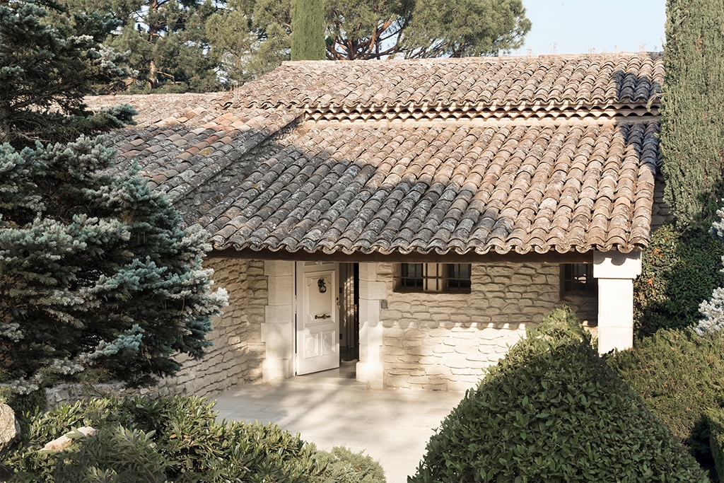 A rustic house with a tiled roof and stone walls, partially obscured by lush green trees and bushes. The front door is slightly open, revealing a glimpse inside. The setting is bright and sunny, highlighting the textures and colors of the building and vegetation.