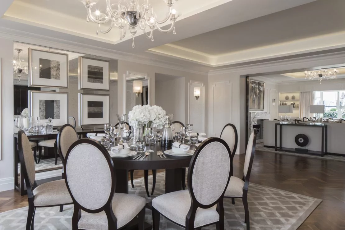 A formal dining room with a round wooden table set for six, reminiscent of Eaton Square elegance. The table is adorned with white flowers in a vase and surrounded by upholstered chairs. The room features refined decor, a chandelier, mirrored walls, and an adjacent open kitchen with contemporary furnishings.