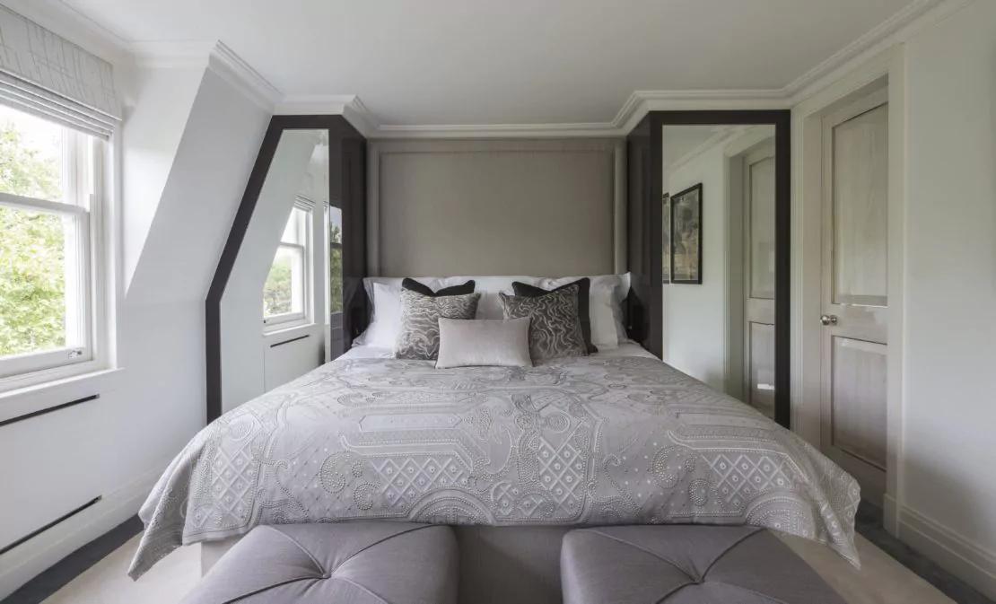 A cozy bedroom in an Eaton Square style, with a large bed centered against the back wall, adorned with patterned gray and white bedding and several plush pillows. The room features two windows on the left, a light gray door on the right, and two gray cushioned benches at the foot of the bed.