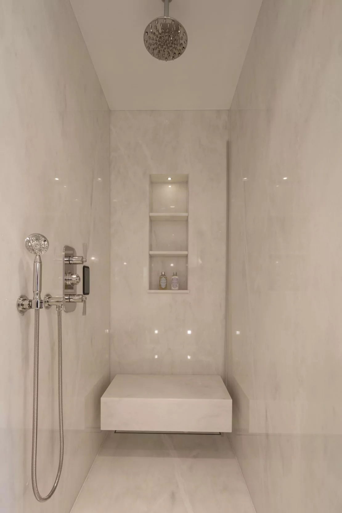 A modern, minimalist shower inspired by Eaton Square, featuring light-colored marble walls and a built-in bench. The shower boasts a large rain showerhead, a handheld showerhead, and a niche with three shelves for toiletries. The overall design is sleek and clean.