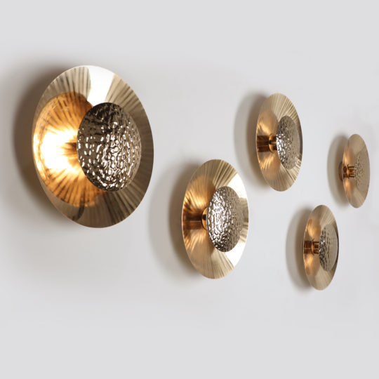 Designer Gold Plated Hammered Round Wall Lights are mounted on a white wall in a staggered arrangement. The lights' gold-plated, disc-shaped design features hammered, textured centers that create a warm, ambient glow with their reflective surfaces.