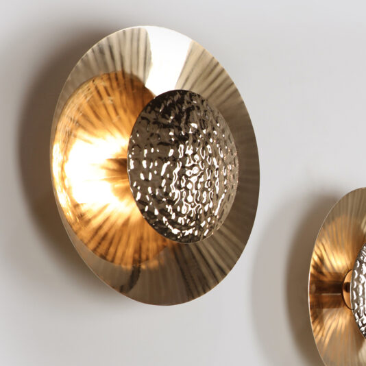 A close-up of two Designer Gold Plated Hammered Round Wall Lights reveals their round, textured surfaces. Each light features a reflective hammered metallic center that emits a warm, glowing light. This enhances the golden tones and adds depth to the pattern against a plain white wall background.