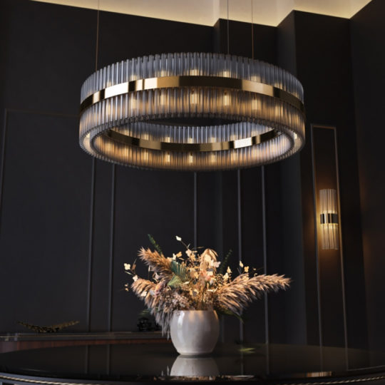A luxurious modern interior showcases the Contemporary Designer Tiered Chandelier adorned with gold accents and intricate metallic tiers. Beneath it, a decorative vase filled with dried flowers is elegantly placed on a glossy black table. The walls feature dark tones complemented by subtle vertical paneling.
