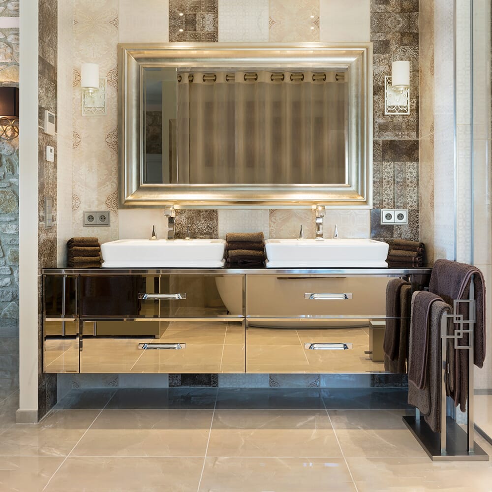 A luxurious bathroom features a double sink vanity with a large framed mirror above. The vanity is set against a wall with intricate tile designs. Two sets of neatly folded brown towels are placed on towel rails. The floor is tiled in a glossy beige finish.