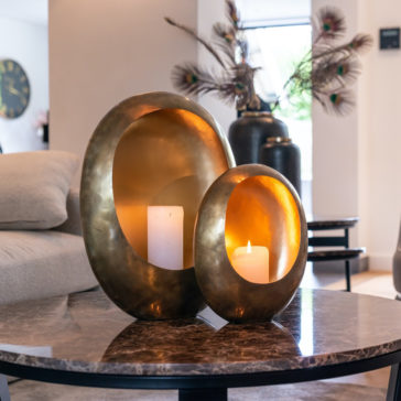 Large Gold Candle Holder Two modern, oval-shaped brass candle holders with white candles sit on a marble coffee table, serving as elegant statement pieces in a stylish living room. One candle is lit, and decorative vases with peacock feathers enhance the chic décor.