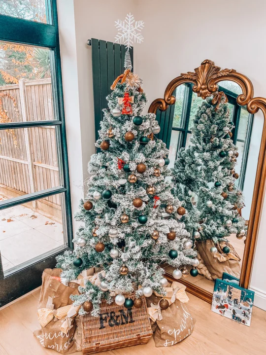 A decorated Christmas tree with a mix of green, brown, and white ornaments stands in front of a large ornate mirror. The tree is topped with a white snowflake. Reflecting in the mirror, this charming piece of Christmas decor is surrounded by wrapped presents and has a basket underneath labeled “F&M".