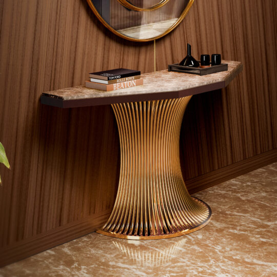 A Marble Demi Console with a curved marble top and gold-plated base stands against a wood-paneled wall, displaying books, a black tray, and decor. A round mirror hangs above, with polished brown marble flooring below.