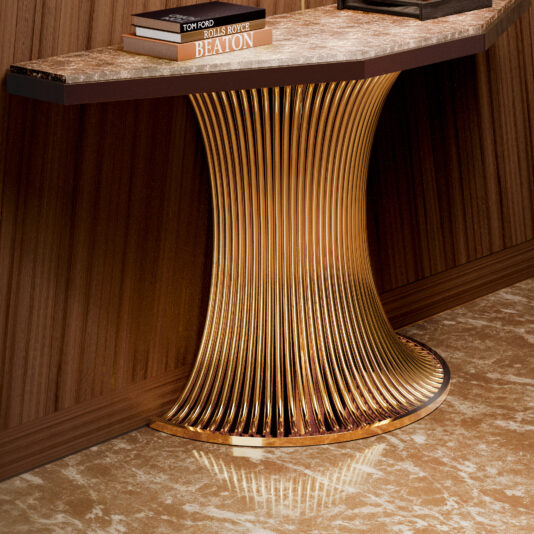 Marble Demi Console The Marble Demi Console features a shiny gold fluted base and dark marble top, standing against a wooden wall. Books are stacked on it, and it rests on a polished brown marble floor.