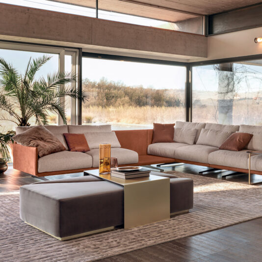 The modern living room has large windows with scenic views and includes a Retro Style Modular Corner Sofa with brown cushions, a square coffee table with glass vases, and a gray ottoman. A tall plant is in the left corner, and the floor features a textured rug.