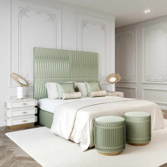 A sophisticated bedroom features a large bed with an elegant sage green headboard. The bed is flanked by Art Deco Style Bedside Cabinets, each adorned with stylish lamps. Two round, sage green ottomans sit at the foot of the bed. The room has ornate white wall paneling.
