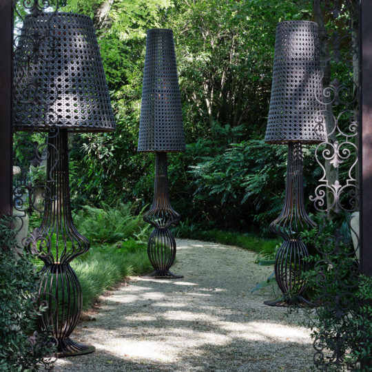 A gravel garden path winds through lush greenery, illuminated by three Large Iron Outdoor Lamps featuring woven shades and curved openwork bases, as sunlight filters through the dense surrounding foliage.