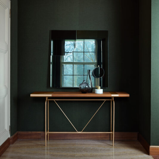 A modern entrance hall features the Modern Leather Console Table adorned with decorative vases and a small round mirror, set against a dark green wall with a geometric mirror reflecting outdoor trees.