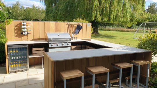 An outdoor kitchen with a stainless steel grill, wooden cabinets, and a mini-fridge embraces the latest garden trend. Shelves with spices and cooking utensils hang above the counter, while bar stools line a countertop in front. A grassy yard and a soccer goal are in the background.