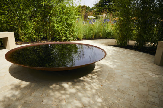 A tranquil, round water feature, surrounded by a lush garden with tall grasses and bamboo plants—a true embodiment of the latest garden trend. The water reflects the sky above and is set within a circular, light-colored stone patio with seating benches on either side.