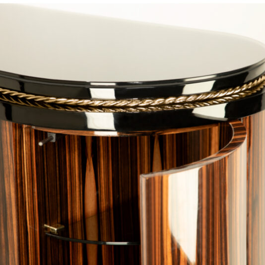 Close-up of the Oval Veneered Sideboard, showcasing its curved glass door. The cabinet boasts a glossy black top, is adorned with a gold braid accent, and displays a striking vertical wood grain pattern. The door is partially open, revealing a glass shelf inside.