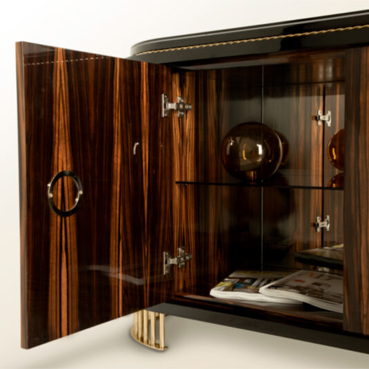 The Oval Veneered Sideboard boasts a high-gloss finish and a distinct grain pattern. One door is open, revealing glass shelves inside. On the top shelf sit two spherical, amber-colored objects, while a stack of magazines rests on the bottom shelf. The oval door handle features a circular design.