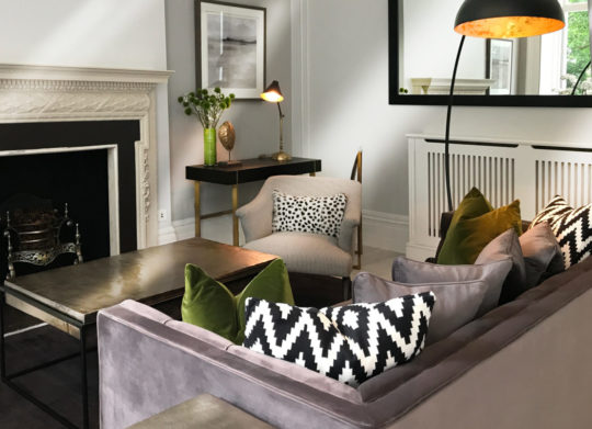 A modern living room featuring a gray sectional sofa adorned with green and black-and-white patterned cushions. A sleek metal coffee table sits in front, adding personality to the space, and a cozy armchair is beside a small desk with a green vase and lamp. A large mirror hangs above a radiator cover.