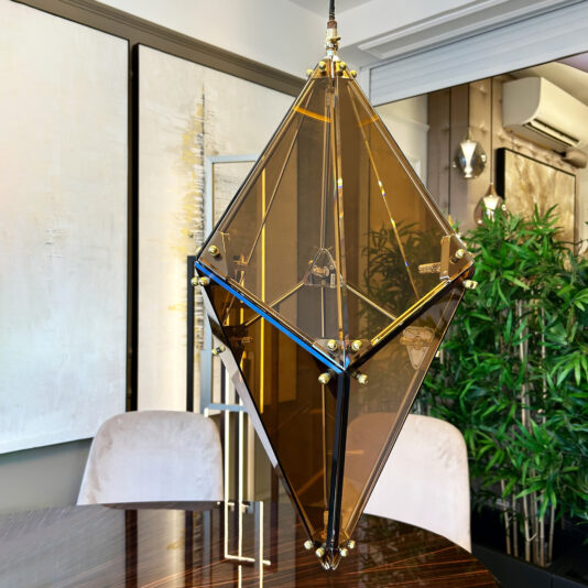 The Modern Champagne Prism Glass Pendant with metallic accents hangs above a glossy wooden table in a modern room adorned with contemporary art and a green potted plant.