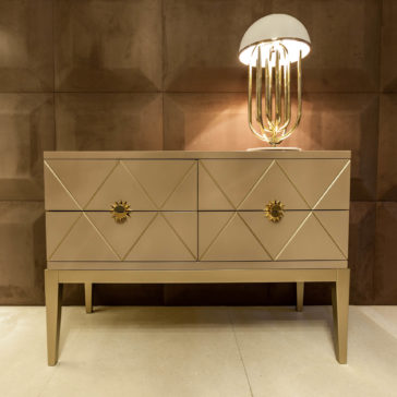 Art Deco Inspired Chest of Drawers The Art Deco Inspired Chest of Drawers in beige features geometric diamond patterns, gold sunburst drawer handles, and a gold lamp with a white dome shade, set against a textured brown wall.