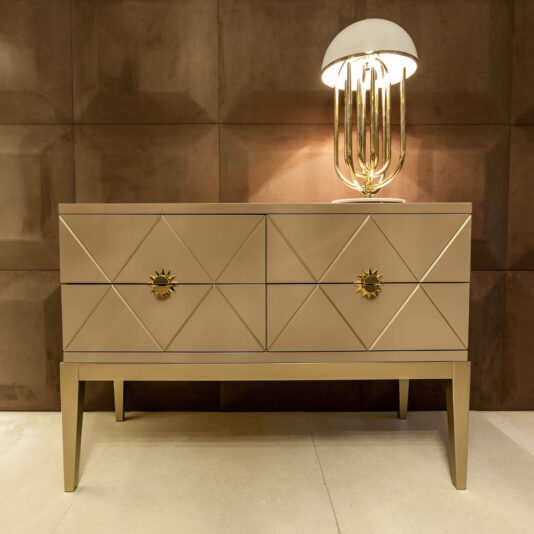 The Art Deco Inspired Chest of Drawers in beige features geometric diamond patterns, gold sunburst drawer handles, and a gold lamp with a white dome shade, set against a textured brown wall.